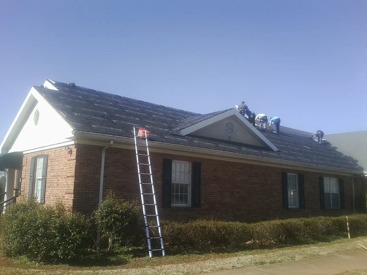 Crew tearing shingles off a brick ranch roof with a ladder set — Emergency Roof Repair work by our crew in Meadow Grove