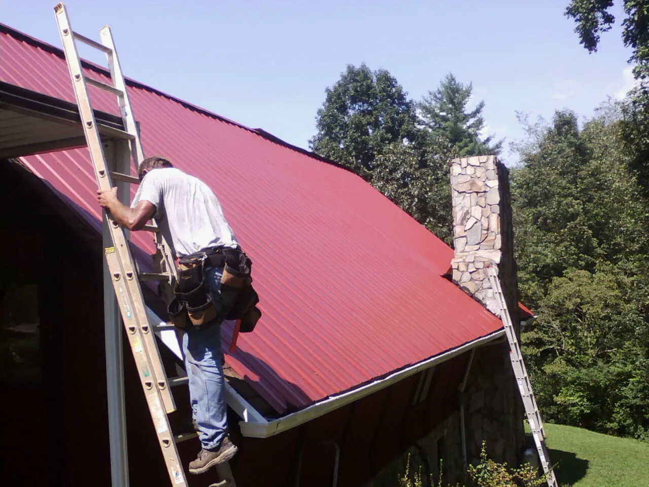 Roofer on a ladder beside a red metal roof and stone chimney — Soffit & Fascia Repair by Roofing Brothers in Meadow Grove